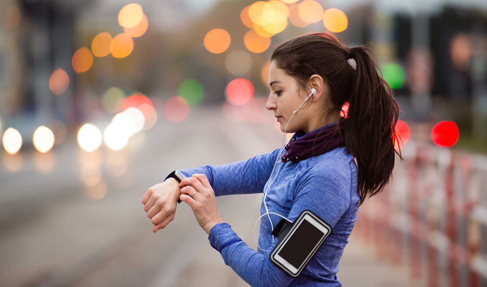 A woman in a blue top checks her fitness tracker, possibly downloaded from free software for personal trainers, while jogging on a city street at dusk, with blurred street lights in the background.